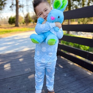 May include: A child wearing light blue pajamas with a white bunny pattern, hugging a blue plush bunny with green and pink accents. The child is also wearing brown slippers. The background features a wooden deck and a blurred green landscape.