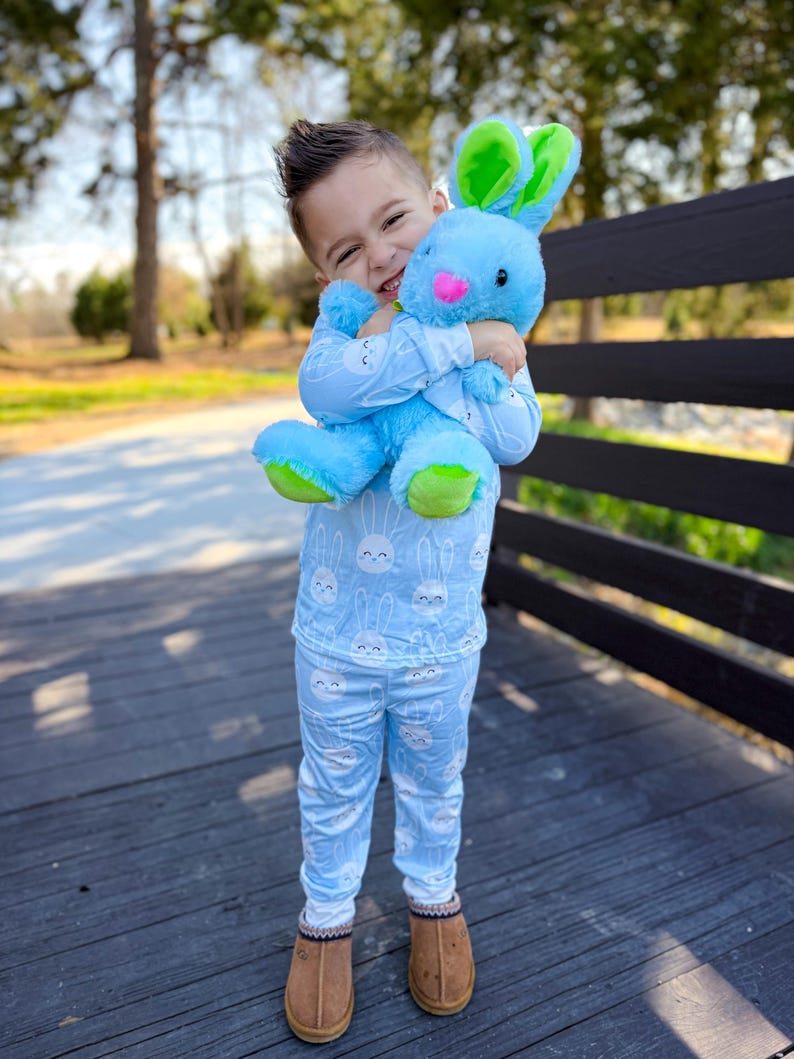 May include: A child wearing light blue pajamas with a white bunny pattern, hugging a blue plush bunny with green and pink accents. The child is also wearing brown slippers. The background features a wooden deck and a blurred green landscape.