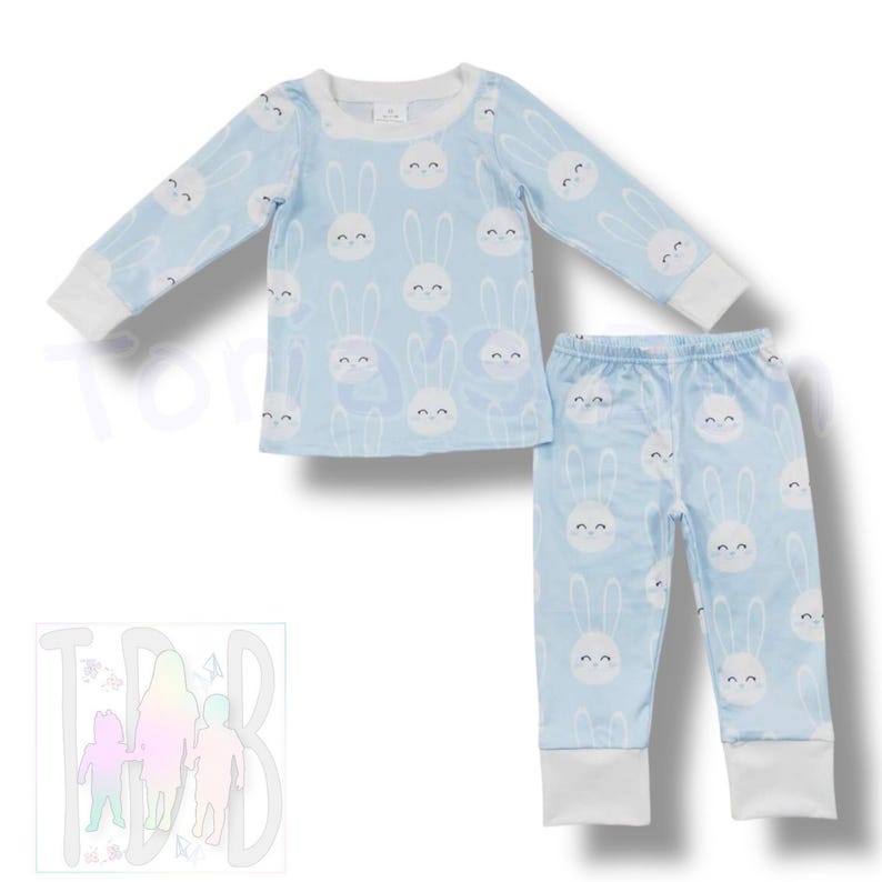 May include: Light blue pajama set featuring a repeating pattern of white bunny faces. The long-sleeved top and pants have white cuffs. The pajama set is designed for children.