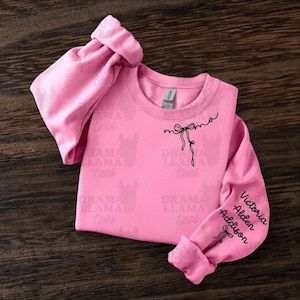 May include: A pink sweatshirt with long sleeves. The word "mama" is written in cursive with a bow graphic. The right sleeve has the names "Victoria, Alden, Addison" in black script. The brand name "Drama Llama Tees" is printed on the shirt.