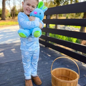 May include: A young child wearing light blue pajamas with a cloud pattern, holding a blue plush bunny with green and pink accents. A brown woven basket sits nearby. The child is wearing brown slippers.