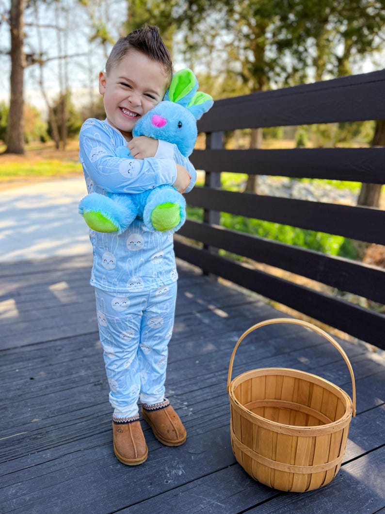May include: A young child wearing light blue pajamas with a cloud pattern, holding a blue plush bunny with green and pink accents. A brown woven basket sits nearby. The child is wearing brown slippers.