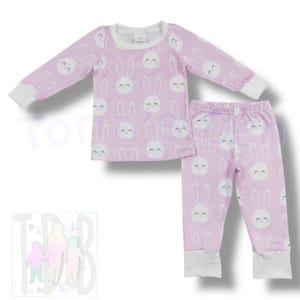 May include: A two-piece pajama set in light purple with a repeating pattern of white bunny faces. The long-sleeved top and pants have white cuffs and trim. The pajamas are designed for children.