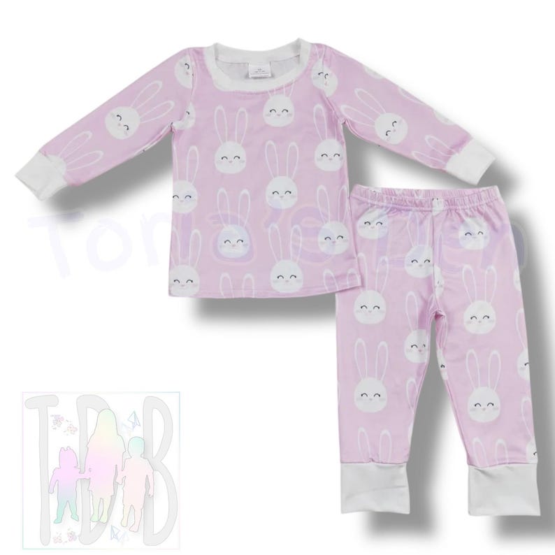 May include: A two-piece pajama set in light purple with a repeating pattern of white bunny faces. The long-sleeved top and pants have white cuffs and trim. The pajamas are designed for children.