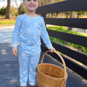 May include: A young child wearing light blue pajamas with a repeating white bunny face pattern. The child is holding a wooden basket with a handle and wearing brown slippers. The background includes a black fence and trees.