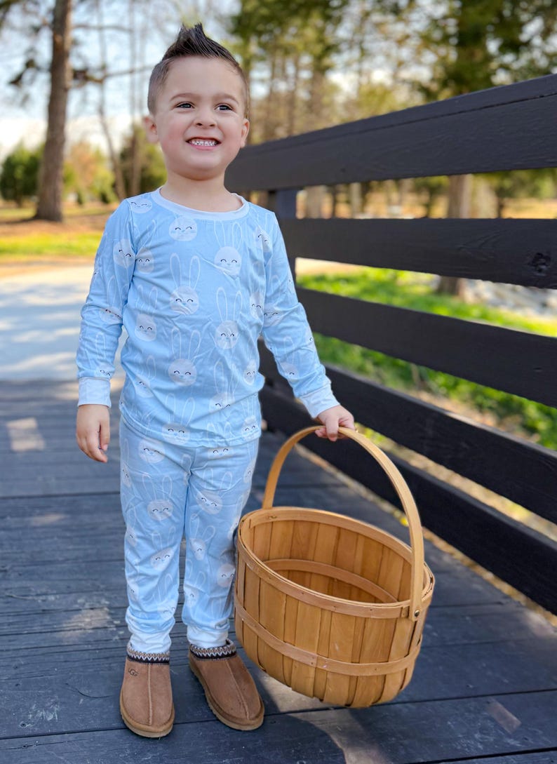 May include: A young child wearing light blue pajamas with a repeating white bunny face pattern. The child is holding a wooden basket with a handle and wearing brown slippers. The background includes a black fence and trees.
