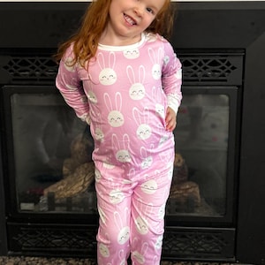 May include: Pink pajama set with a white bunny face pattern. The long-sleeved top and pants have white trim. The child is smiling and standing in front of a fireplace.