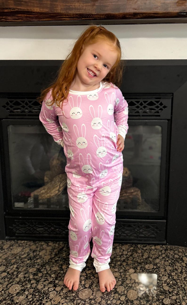 May include: Pink pajama set with a white bunny face pattern. The long-sleeved top and pants have white trim. The child is smiling and standing in front of a fireplace.
