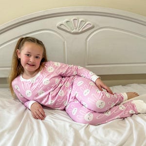 May include: Pink pajama set with a white bunny face pattern. The long-sleeved top and pants have white trim at the cuffs and neckline. The pajamas are worn by a person lying on a white bed.