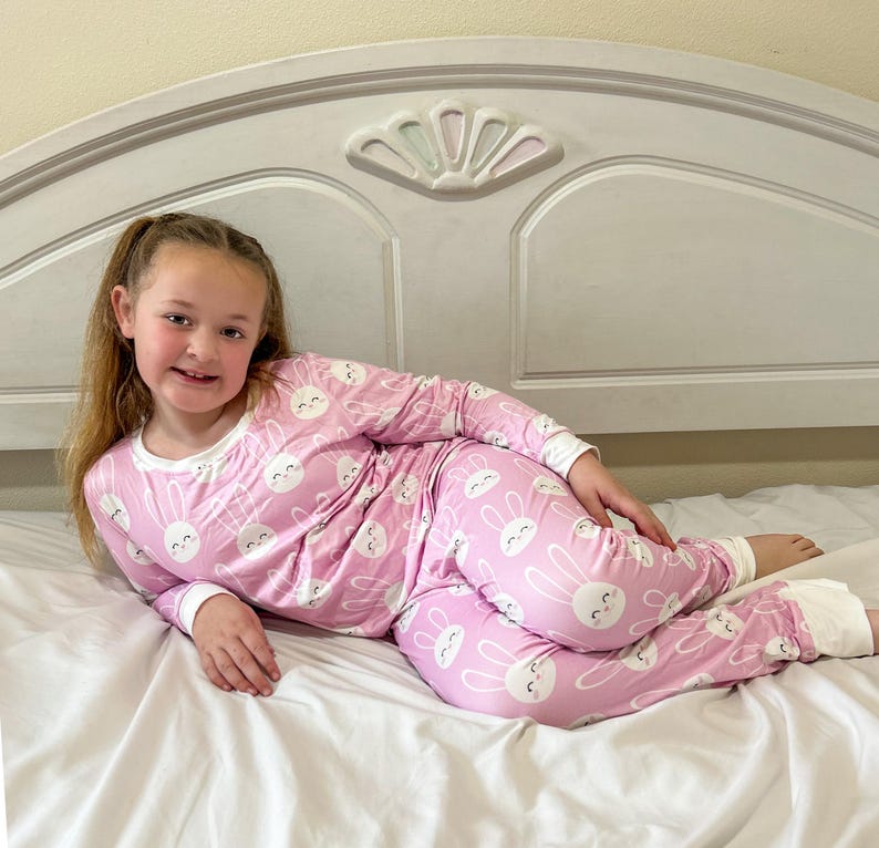 May include: Pink pajama set with a white bunny face pattern. The long-sleeved top and pants have white trim at the cuffs and neckline. The pajamas are worn by a person lying on a white bed.