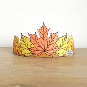 May include: A paper crown featuring autumn leaves in shades of orange and yellow. The crown has a brown base and includes the text "HAPPY PAPER TIME" on a small white tag. The leaves are detailed with black veins.