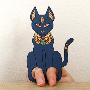 May include: Blue finger puppet of an Egyptian cat with a gold collar and a scarab beetle. The cat has red eyes and a gold ankh symbol on its forehead.