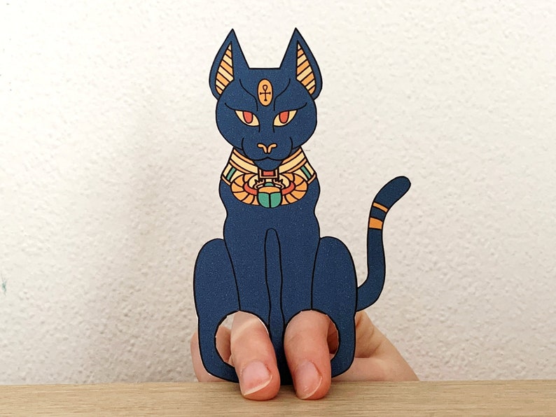 May include: Blue finger puppet of an Egyptian cat with a gold collar and a scarab beetle. The cat has red eyes and a gold ankh symbol on its forehead.