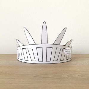 May include: A white paper crown featuring a pointed, radiating design reminiscent of the Statue of Liberty. The band is decorated with a series of rectangular outlines. A small circular logo with the text HAPPY PAPER TIME is visible.