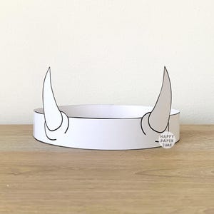 Pode incluir: Uma coroa de papel branco com dois chifres curvos, delineados em preto. A coroa tem uma faixa circular e o texto "HAPPY PAPER TIME" em uma pequena etiqueta. A coroa está sobre uma superfície de madeira.