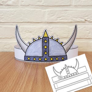 May include: A paper Viking helmet crown featuring a grey dome, large curved horns, and a dark blue band with yellow circular accents. The design includes a central vertical strip and a matching black and white printable template sheet below it.