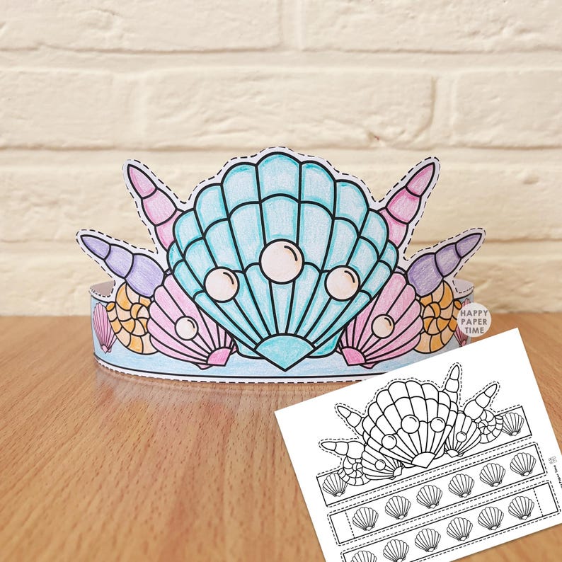 Pu&ograve; includere: Una corona di carta con una grande conchiglia blu centrale con tre perle bianche, affiancata da conchiglie pi&ugrave; piccole rosa, viola e arancioni. Un foglio modello stampabile in bianco e nero con contorni di conchiglie si trova in primo piano.