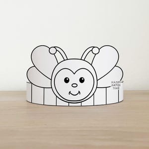 Può includere: Una corona di carta in bianco e nero con un design a forma di ape, dotata di un volto sorridente, antenne e ali. La fascia presenta un motivo a righe verticali e il testo HAPPY PAPER TIME è stampato in inchiostro nero sull'ala destra dell'ape.