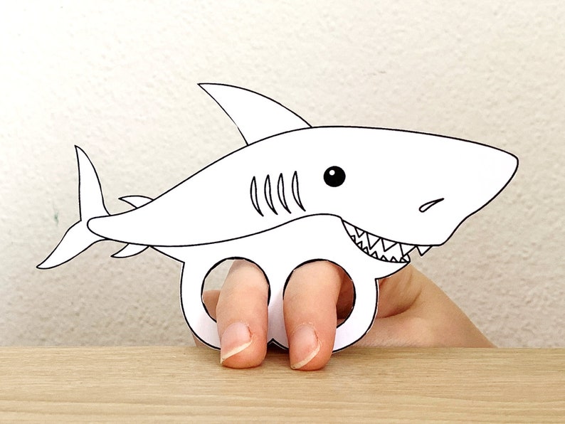 May include: A white paper shark finger puppet with a black outline and a toothy grin. The shark has a black eye and gills.