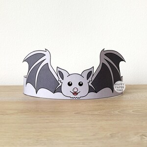 May include: A paper bat crown with a gray bat design. The bat has large wings and a smiling face with a pink nose and tongue. The crown is white with the text "HAPPY PAPER TIME" on the side.