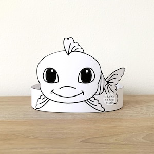 May include: A black and white paper fish crown with a smiling face and the text "HAPPY PAPER TIME". The crown is designed to be worn and is suitable for children's crafts and activities.