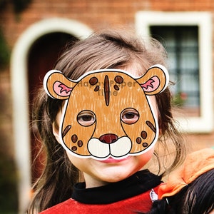 May include: A child wearing a brown and white paper mask of a cheetah. The mask has pink ears and black spots. The child is smiling.