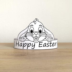 May include: A white paper Easter headband with a cartoon bunny design. The headband displays the words "Happy Easter" in black. The bunny has large eyes and long ears, perfect for Easter celebrations.