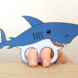 May include: Blue and white paper shark finger puppet with a toothy grin. The shark has a black eye and a white belly. The puppet is designed to fit on a finger.