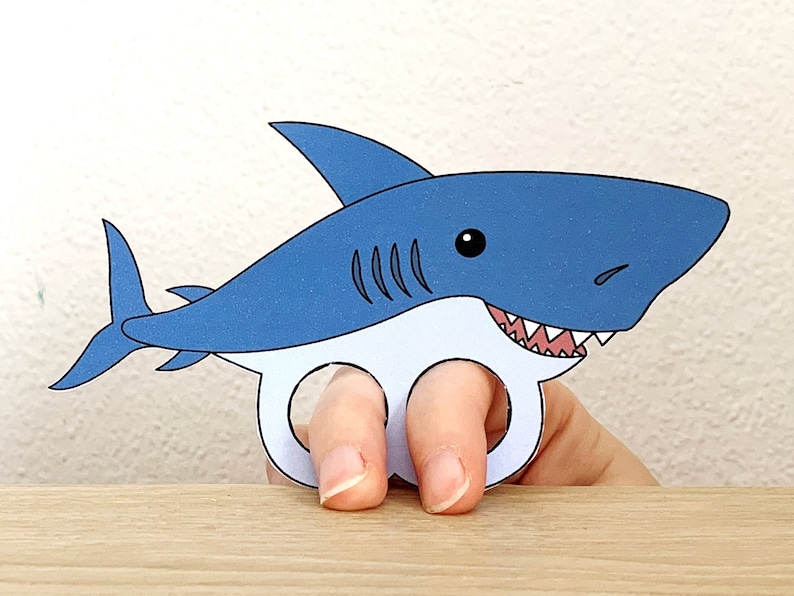 May include: Blue and white paper shark finger puppet with a toothy grin. The shark has a black eye and a white belly. The puppet is designed to fit on a finger.