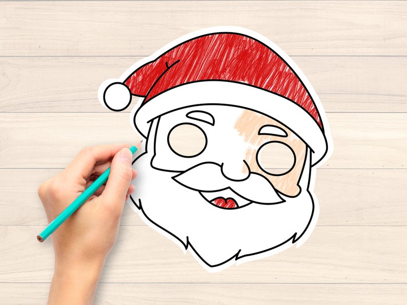 May include: A hand with a teal pencil is colouring a Santa Claus face outline. The Santa face has a red hat, white beard, and a smiling mouth. The background is a light wood grain.