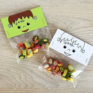 May include: Two clear plastic treat bags with green and white Frankenstein faces printed on the top. The bags are filled with colorful candy.