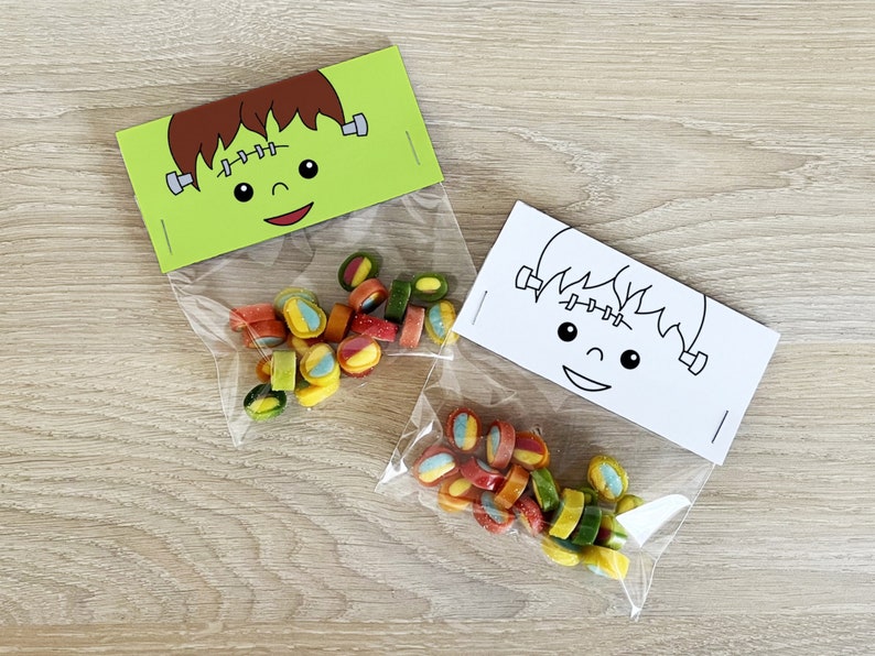 May include: Two clear plastic treat bags with green and white Frankenstein faces printed on the top. The bags are filled with colorful candy.