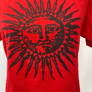 May include: Red t-shirt with a black graphic of a sun with a face.