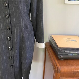 May include: A grey pinstripe dress with a white collar and cuffs. The dress has a button-up front and a fitted silhouette.