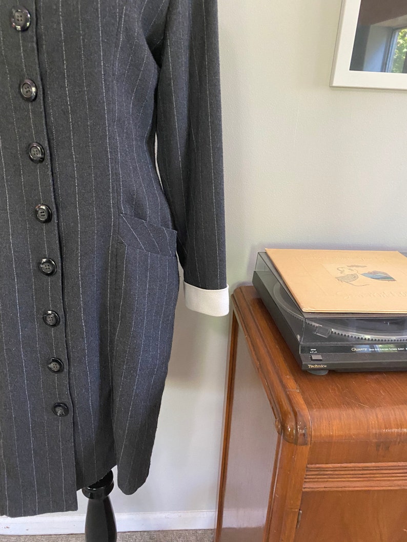 May include: A grey pinstripe dress with a white collar and cuffs. The dress has a button-up front and a fitted silhouette.