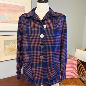 May include: A blue and rust plaid jacket with large white buttons and two front pockets. The jacket is displayed on a mannequin. The jacket has a collared neckline and long sleeves. The plaid pattern is a classic design.