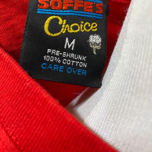 May include: A black and white tag with blue stripes and the text "Soffe's Choice" in yellow. The tag also says "M", "Pre-Shrunk 100% Cotton", and "Care Over".