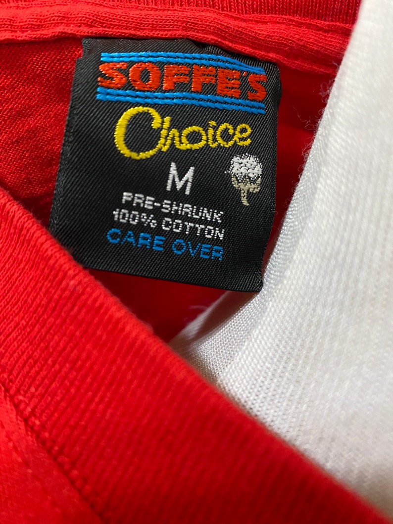 May include: A black and white tag with blue stripes and the text "Soffe's Choice" in yellow. The tag also says "M", "Pre-Shrunk 100% Cotton", and "Care Over".