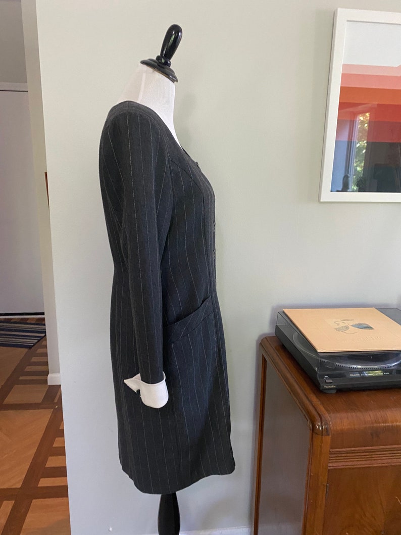 May include: A grey pinstripe dress with a white collar and cuffs. The dress has a button-up front and a pocket on the left side.