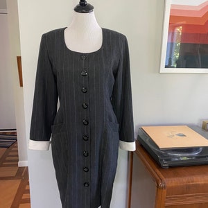 May include: A grey and white striped dress with a button-up front and white cuffs. The dress has a fitted silhouette and pockets on the sides.