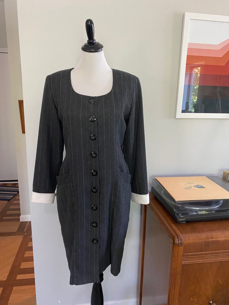 May include: A grey and white striped dress with a button-up front and white cuffs. The dress has a fitted silhouette and pockets on the sides.