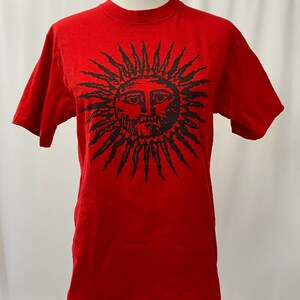 May include: Red t-shirt with a black sun graphic featuring a face. The sun has a spiky outline.