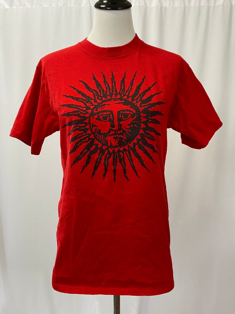 May include: Red t-shirt with a black sun graphic featuring a face. The sun has a spiky outline.