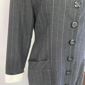May include: A long-sleeved, grey pinstripe dress with a white collar and white cuffs. The dress has a button-up front and a single pocket on the left side.