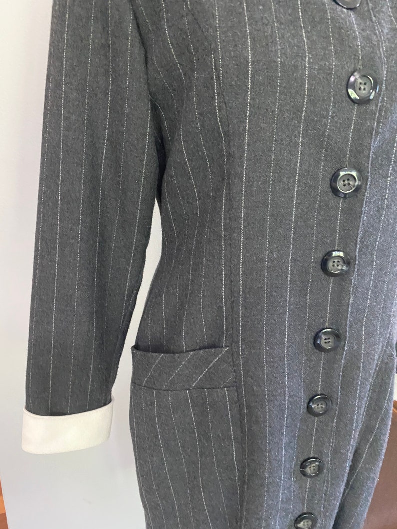 May include: A long-sleeved, grey pinstripe dress with a white collar and white cuffs. The dress has a button-up front and a single pocket on the left side.