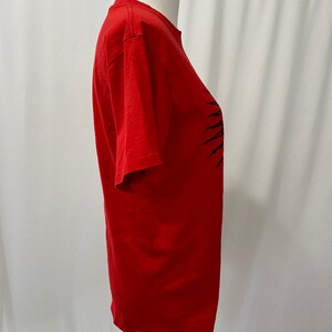 May include: A red short-sleeved t-shirt with a black graphic design of a sun with rays. The graphic design is on the right side of the shirt.