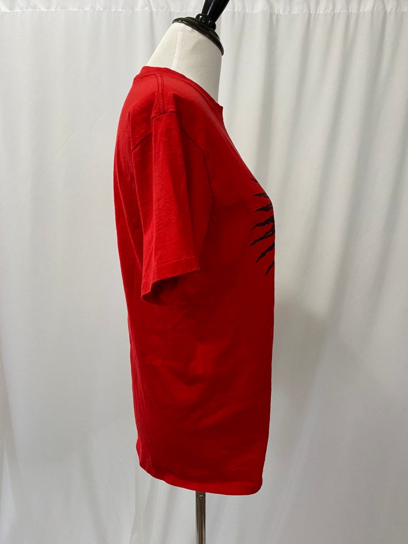 May include: A red short-sleeved t-shirt with a black graphic design of a sun with rays. The graphic design is on the right side of the shirt.