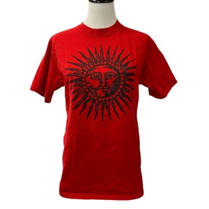 May include: Red short-sleeved t-shirt with a black sun graphic featuring a face.