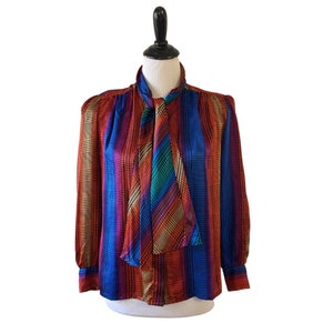May include: A long-sleeved blouse with a colorful geometric pattern in shades of red, blue, green, and gold. The blouse has a tie neck and a button closure.