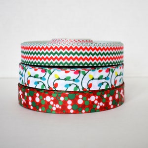 May include: A stack of three Christmas-themed ribbon spools. The top spool has a red and green chevron pattern, the middle spool has a white background with colorful Christmas lights, and the bottom spool has a red and green polka dot pattern.
